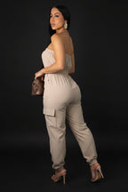 Woman wearing a beige jumpsuit holding a brown handbag on a gray background