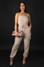 Woman wearing a beige strapless jumpsuit holding a clutch against a gray background