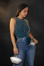 Woman wearing a green sleeveless top with white polka dots and blue jeans, holding a white handbag on a gray background.