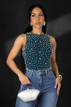 Woman wearing a teal sleeveless top with white embellishments and blue jeans, holding a white handbag on a gray background.