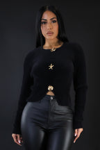 Woman wearing a black sweater with gold buttons and black pants against a plain background
