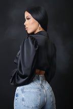 Woman wearing a black satin blouse and light blue jeans against a gray concrete wall.